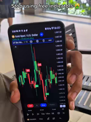 A person holds a smartphone displaying a trading app with a candlestick chart. The phone is held in front of a blurred background, possibly a public space. The app shows green and red candlesticks, with buy and sell signals. The top of the image has text overlayed.