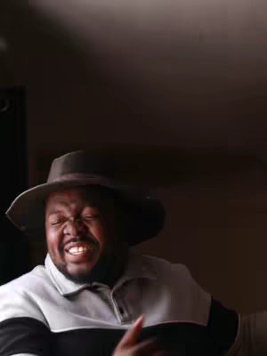 The image shows a man smiling with his eyes closed. He wears a hat and a polo shirt, and the background is a simple, dark wall.