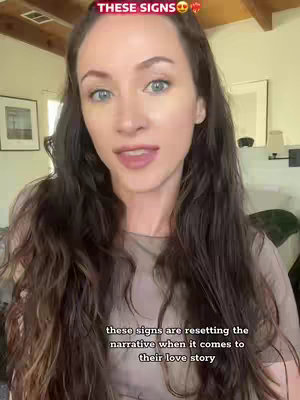 A woman with long brown hair poses looking toward the camera, slightly smiling. The background includes light-colored walls and wood accents, with a hint of a doorway. The text overlay at the top reads 'THESE SIGNS' with a heart emoji.