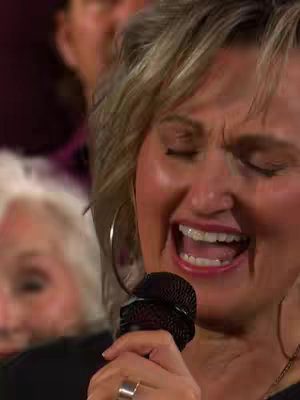 The cover image features a close-up of a woman singing with her eyes closed and mouth wide open, holding a microphone. Her blonde hair is styled, and she wears large hoop earrings and a ring. In the background, slightly out of focus, are other people.