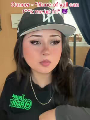 The image is a close-up selfie featuring a young woman. She is wearing a black baseball cap with a white letter on it, and a black top with a green logo. She has long dark hair and is looking slightly to the side with a neutral expression. Above her, a text overlay reads 'Cancer - "None of yall can f**k me up fr!"' followed by a devil emoji.