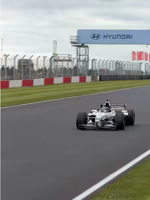 An F1 race car speeds around a racetrack. The car is silver-grey with large black tires, positioned on the right side of the frame. The track is dark grey, bordered by a green grassy area and red-and-white striped barriers. In the background, there is a large sign that reads "HYUNDAI". The sky is overcast and grey.