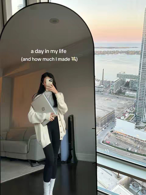 A woman stands in front of a large, arched mirror, holding a laptop. The mirror reflects the woman and the interior of a room, while a large window to the right reveals a city skyline at sunset. The woman is wearing a white cardigan over a light colored top, black leggings, and white socks. She is in a well lit interior with a window that offers a view of city buildings and the sky.