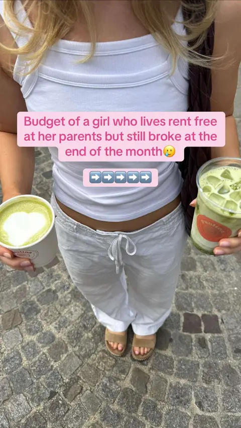 A woman in a white tank top and off-white drawstring pants stands on a cobblestone street, holding a matcha latte in one hand and a green iced drink in the other with two more drinks in front of her. A pink text overlay at the top says, "Budget of a girl who lives rent free at her parents but still broke at the end of the month🥲 " with arrow emojis.