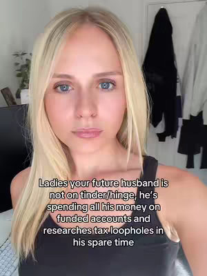 A woman with blonde hair is centered in a selfie-style video shot. She has fair skin, blue eyes, and is looking directly at the camera with a neutral expression. She's wearing a black tank top. Behind her, a wall, a door, and the edge of some objects are visible, but the background is slightly out of focus.