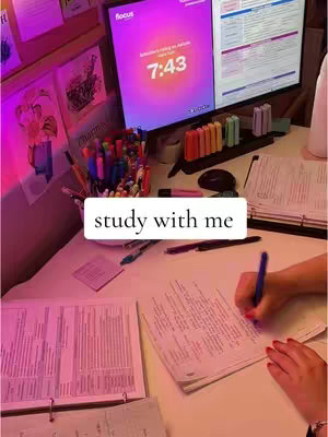 A desk with papers, pens, markers, and monitors. The overall tone is pink and well-lit. A hand is writing on paper.