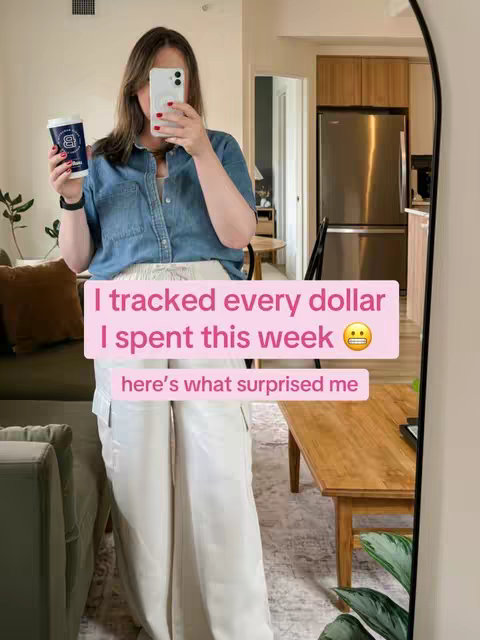A woman in a blue denim shirt and off-white pants stands in front of a mirror, holding a coffee cup and a phone. The image is brightly lit, with neutral colors and a clean, modern interior visible.