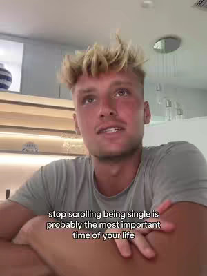 A young man with spiky blond hair is looking up and speaking to the camera. He is wearing a gray t-shirt. The background is a light-colored room with some shelves and a light fixture.