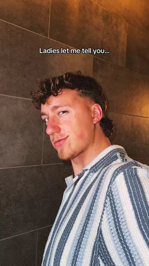 A medium shot features a young man with short, curly brown hair and a slight smirk. He wears a blue and white striped shirt. The background is a textured wall in shades of brown and gray. Above the man's head, text overlayed in the image reads 'Ladies let me tell you...'