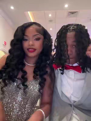 A photograph captures a couple, likely at a formal event, as a woman with elaborate curly black hair and sparkly dress smiles while looking at the camera next to a man in a grey suit and red bow tie.