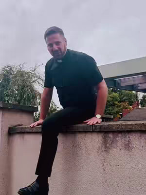 A man in a priest's collar and black shirt is sitting on a concrete wall. The background is a cloudy sky and some greenery.