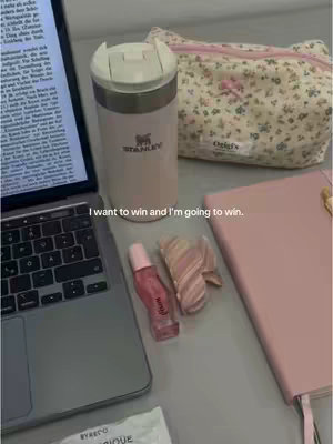A study desk is shown with a laptop, a pink insulated tumbler, a floral patterned cosmetic bag, a pink journal, a tube of makeup, and pink hair clips. Text overlayed on the image says: 'I want to win and I'm going to win.'