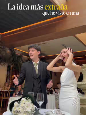 A candid shot of a wedding reception. A man in a suit has his arms out, looking surprised, while a woman in a white dress covers her face with her hands, looking shocked. The setting appears to be a wedding reception.