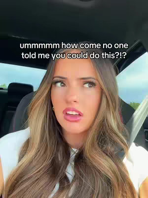 A young woman with long, light brown hair is seated in the front seat of a car, looking toward the right with a surprised or questioning expression. Her lips are slightly parted, suggesting she may be speaking. The car's interior appears dark, with a hint of the outside world visible through the window in the background. The lighting in the image is neutral-toned, with the woman's face brightly lit.