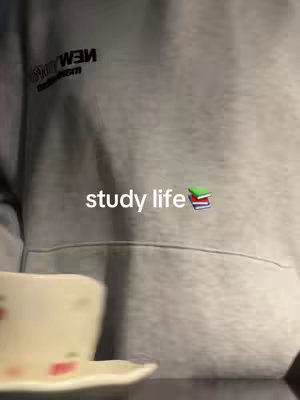 The image shows a close-up of a person wearing a gray hoodie. The text "study life" is overlaid on the image, along with stack of books emoji. In the foreground, a white teacup rests on a saucer.