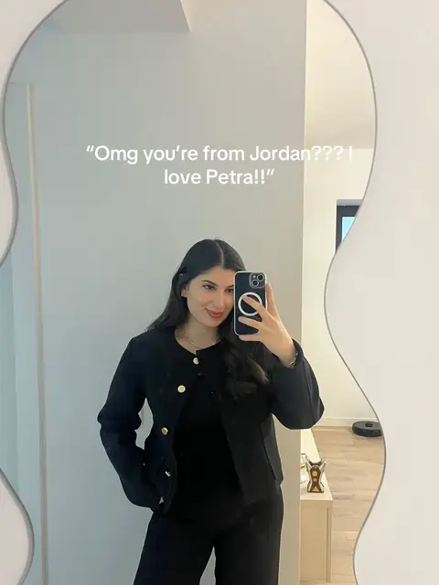 A woman in a black suit is taking a mirror selfie. In the mirror reflection, a room is visible, including a grey wall and a modern curved mirror. The selfie displays text overlaid. The woman appears to be smiling and holding a phone.