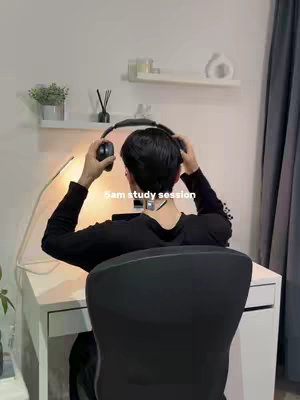 A person is seen from behind, wearing a black long-sleeved shirt and headphones, seated at a white desk and turning towards the right side. The background details a white wall with a shelf holding candles and other decorative items, with a small plant in a pot. The room has soft lighting, and a desk lamp is positioned on the left side of the desk. The text '5am study session' is displayed across the image.