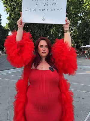 A woman in a vibrant red outfit stands holding a white sign above her head. She has red hair and fair skin. Her outfit includes a strapless red dress and a large, fluffy red boa-like jacket. The background is a blurred outdoor scene, possibly a plaza, with trees and buildings visible.