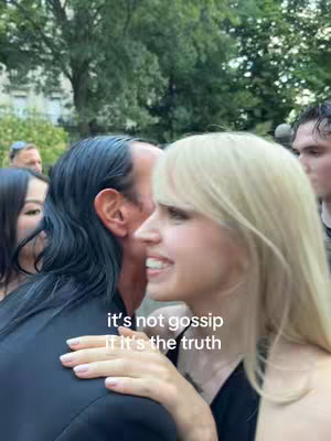 A blonde woman with a light complexion smiles candidly while embracing a man with dark hair. The man is mostly obscured from view. The background is a lush outdoor scene.