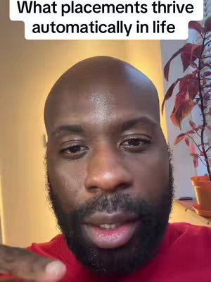 A man with a bald head and a full dark beard is centered in the frame, looking towards the camera. Above his head, text reading "What placements thrive automatically in life" is visible. The background includes a wall and a glimpse of foliage.