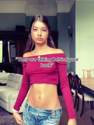 A young woman stands in front of a blurred interior background. She wears a dark red off-the-shoulder long-sleeved crop top and blue denim low-waisted pants. The text overlay says, "They are talking behind your back".