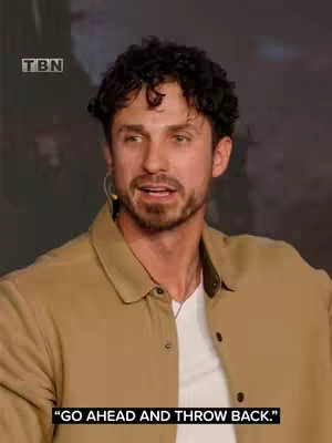 The image features a man with curly dark hair and a trimmed beard against a blurred background. He is wearing a tan button-down shirt over a white t-shirt, with a microphone attached to his ear. Text within a black box at the bottom reads "GO AHEAD AND THROW BACK."