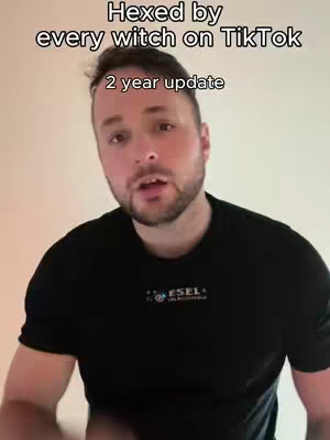 A man with short brown hair and a beard is centered in the frame, looking towards the camera with an open mouth. The background is a plain, light-colored wall. The man is wearing a black t-shirt. White text overlays the top, and a smaller text appears beneath it.