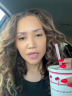 A woman with long, wavy brown hair is seated in a car, facing the camera. She has a neutral expression, with her lips slightly parted as if speaking. She is holding a white cup with red branding and a straw with her right hand, showcasing bright red nail polish. The car’s interior is visible, including a window with sunlight. The background appears blurred.