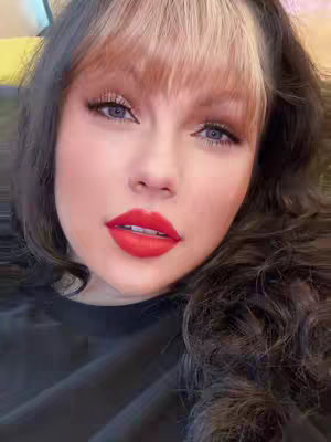 A close-up selfie features a woman with pale skin, striking red lipstick, and a dark top. Her brown hair, styled with a blonde highlight in the front, frames her face. The background is blurred, keeping all focus on her face.