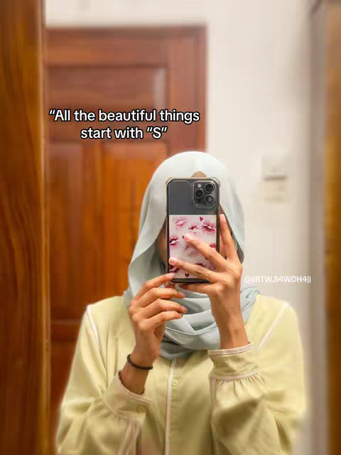 A person is taking a mirror selfie while wearing a light yellow shirt and a light blue headscarf. The person is holding up a phone to cover up most of their face. The text overlay at the top says, "All the beautiful things start with 'S'". Behind the person is a brown wooden door and wall. The image has a soft, warm lighting.