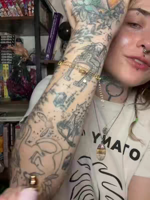A person with arm tattoos and a white shirt is holding a product. The background is a room with shelves, books, and statuettes.