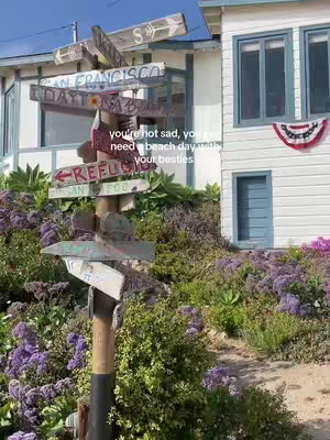 The image presents a sunny outdoor scene featuring a wooden signpost with multiple directional signs pointing to various locations, set against a backdrop of a white house with blue-trimmed windows and vibrant purple flowers. Text overlays are added to the image.