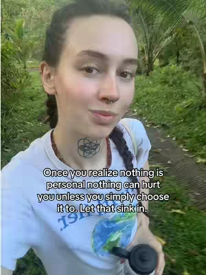 A woman is shown from the chest up in a selfie-style shot taken outside. She's wearing a white t-shirt featuring a graphic of the Earth, and she has a small tattoo on her neck. She is holding a dark-colored object, possibly a bottle or a microphone. The background consists of lush, green vegetation and a walking path, suggesting an outdoor setting. The lighting appears natural and soft. Text is added as a hook in a white font.