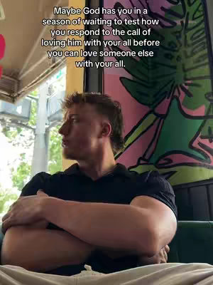 A man with short, styled hair sits in a cafe setting, his arms crossed, looking away from the camera with a thoughtful expression. The background includes a partial view of a colorful mural and window, hinting at an indoor/outdoor space. The lighting is soft and natural.