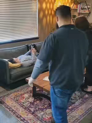 The image shows a room with a couch, a Persian rug, and large windows. An individual is laying on the couch, possibly deceased, with two others standing by. A man in a dark top and jeans is in the foreground, and a woman is to the right with her back turned. There is a person on the couch with someone checking for vitals. The scene implies a death setting.