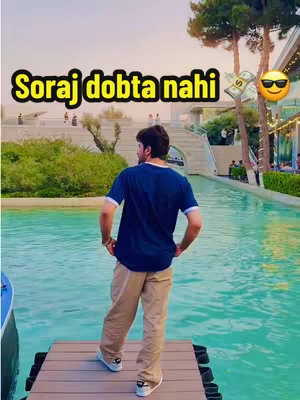 A man stands on a wooden dock with his back to the camera, overlooking a still body of turquoise water. The background includes lush greenery, a bridge, and parts of buildings, suggesting an outdoor location. The sky is visible above, and the overall lighting suggests a bright, sunny day. Text overlay is in yellow, positioned at the top middle of the image.