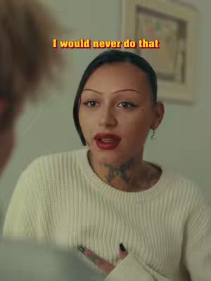 A woman with dark hair and tattoos looks slightly to the side, facing another person out of frame. Text in a yellow and red outline appears above her head. She is wearing a cream-colored sweater and red lipstick.