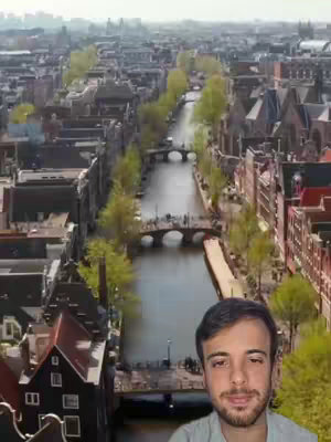 An aerial view of a canal in Amsterdam, with buildings lining both sides and bridges crossing the water. The image includes a man in the foreground, placed over the bottom right quadrant, partially obstructing the view of the buildings and canals. The overall vibe is sunny, suggesting a day of exploration.