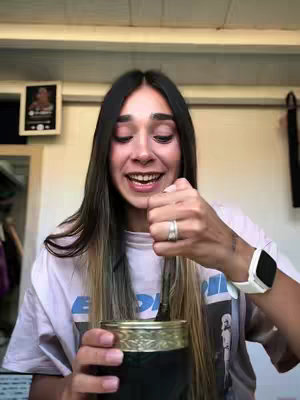A woman with long, dark hair with blonde highlights, looks directly at the camera with a smile. She is stirring a drink in a cup with a metal straw and has a smartwatch on her wrist. The background is a neutral-toned room with a picture on the wall.