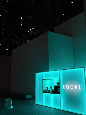 An image of a modern, illuminated structure with a turquoise glow. The structure is composed of rectangular panels, possibly made of glass or a similar translucent material, with a clearly labeled opening at a lower level, which is where the word LOCAL is projected. The background is dark.