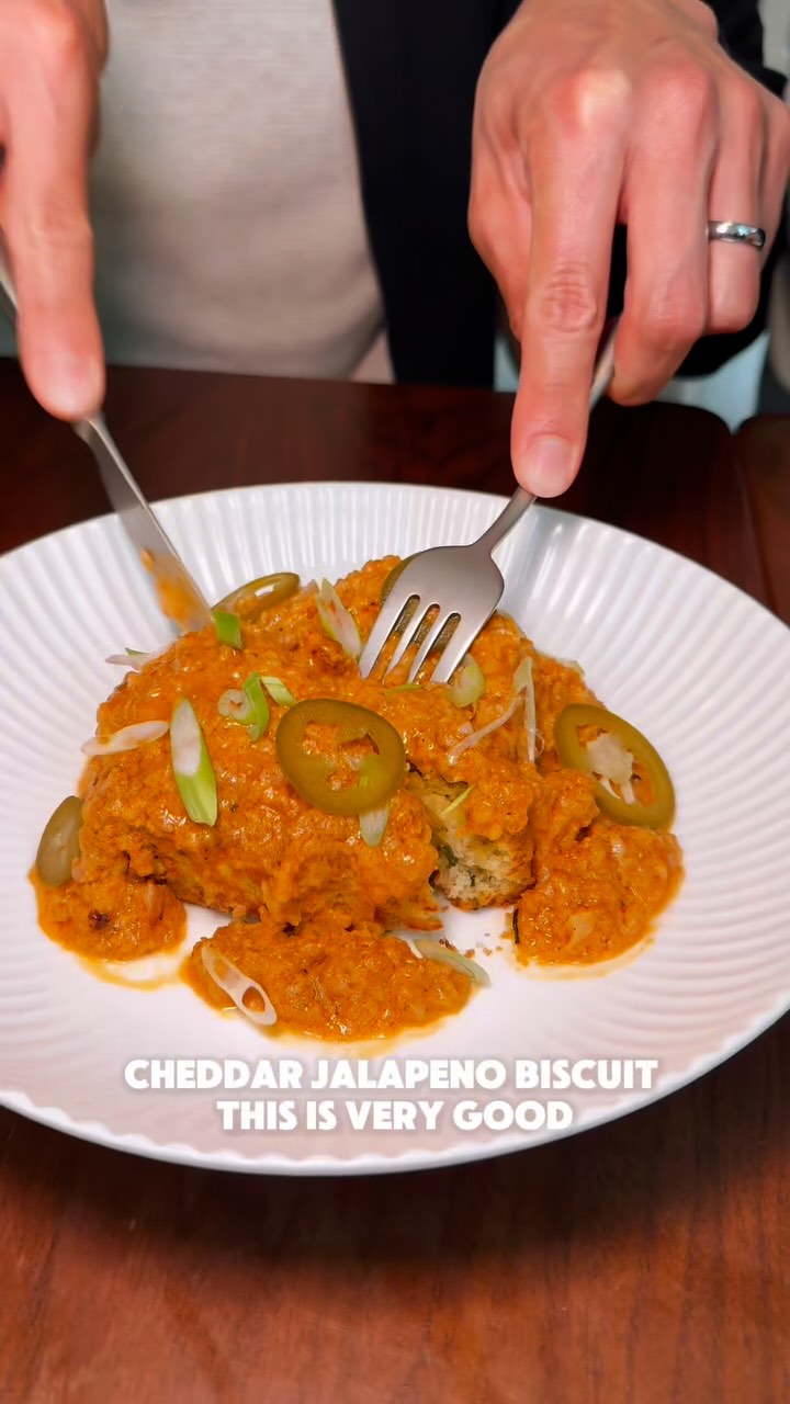 The image captures a close-up shot of food being served on a white plate. The main focus is a biscuit covered with a bright orange sauce, garnished with green onions and slices of jalapeño. A fork and knife are in the frame, indicating someone is about to cut into the dish. The plate rests on a dark wood surface, with the background slightly blurred.