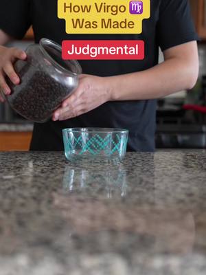 A person in a black shirt is pouring dark beans from a large glass jar into a clear glass bowl with a teal geometric design. The scene is set on a countertop with a speckled gray surface. Text overlays at the top read 'How Virgo Was Made' in yellow, with the Virgo symbol, and 'Judgmental' in red.