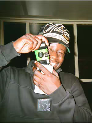A smiling young man in a black jacket and a cap is holding a green and white camera in front of his face. He is standing in front of a window.