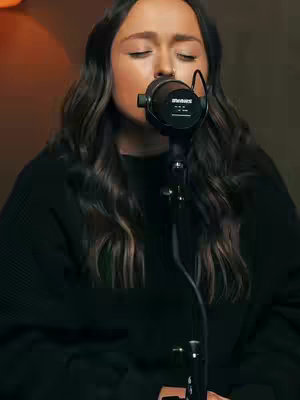 Close-up shot of a woman singing into a microphone, eyes closed. Her long, dark hair frames her face, which conveys a sense of emotion and focus. The microphone is the central element, and the background is a warm, blurred color.