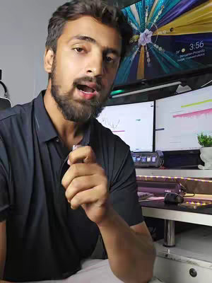 A man with a beard is speaking to the camera. He is holding a small object and has multiple computer monitors behind him displaying charts and graphs. The background includes a desk with items and a clock displaying the time.