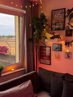 A cozy bedroom shot with an open window revealing an outdoor view. The room is lit with warm fairy lights, draped curtains, and dark-colored furniture. Decorations include framed pictures and a plant with a prominent display.