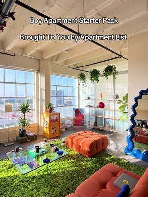 An eye-level shot of a stylish, modern apartment interior. The focal point is a large green shag rug covering the floor, with an orange ottoman in the center. A clear glass coffee table sits on the rug, displaying various items. Large windows let in bright sunlight. Walls are white. The overall aesthetic blends contemporary and minimalist elements with a pop of color.