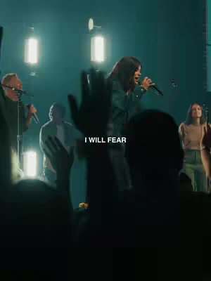 A live stage shot with stage lights illuminating a woman singing into a microphone, with silhouetted audience members in the foreground raising their hands. Text overlayed reads 'I WILL FEAR'.