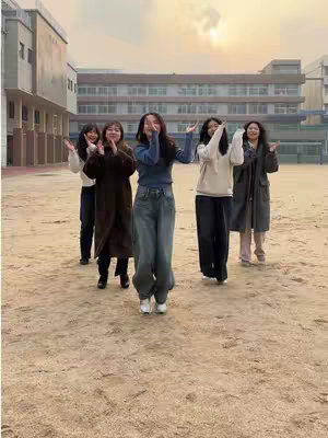 Five young women are standing in an outdoor setting, possibly a schoolyard or park, with a large brick building in the background. They are all smiling and have their arms raised in the air, suggesting a gesture of praise or joy. The lighting indicates it is daytime, and the overall feel is positive, energetic, and celebratory.