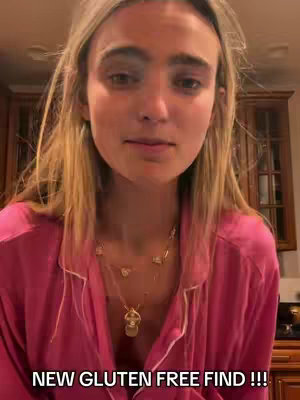 A woman with blonde hair is looking at the camera. She is wearing a pink shirt and a necklace. Below her, in large white font, is the text: NEW GLUTEN FREE FIND !!! Background shows a kitchen setting with warm lighting and wooden cabinets.